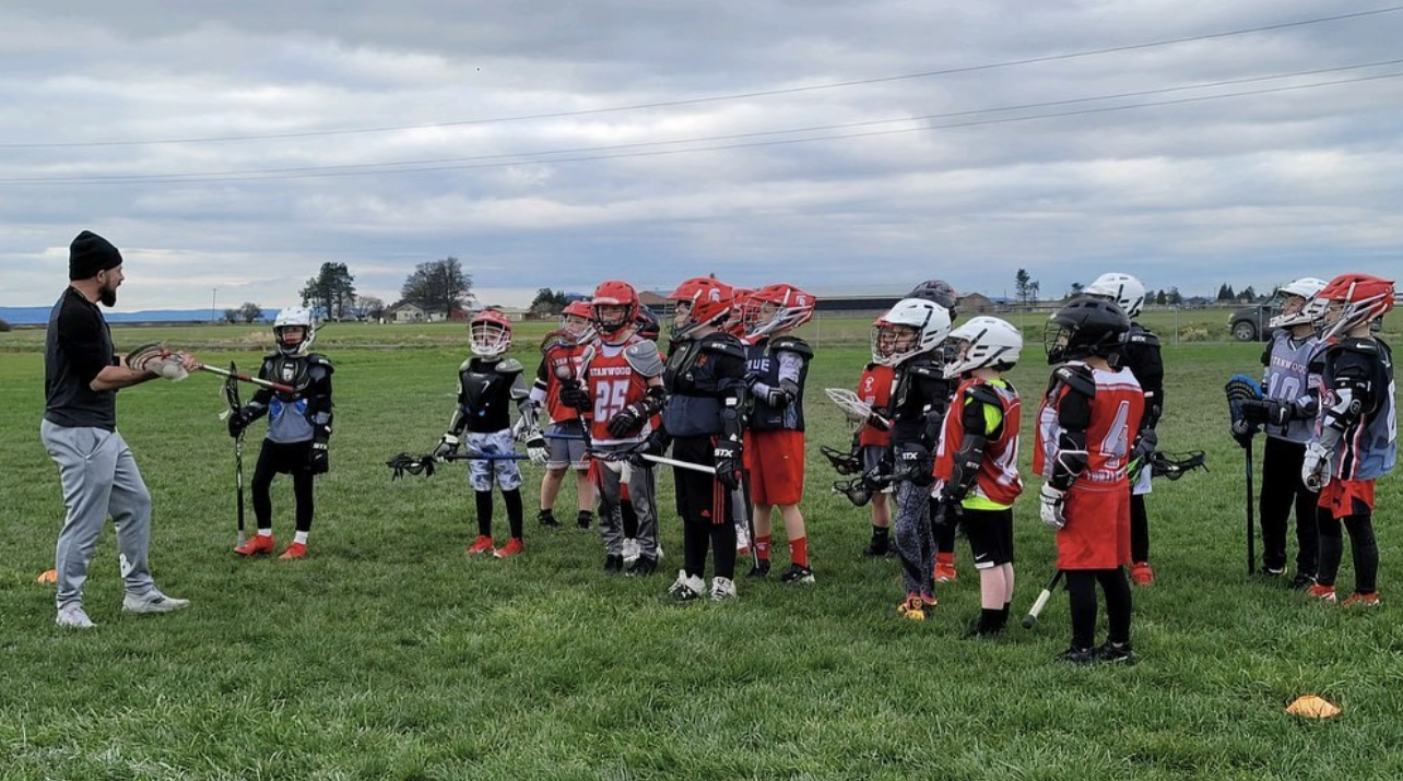 A coach instructing a youth lacrosse group standing in a line on an open grass field