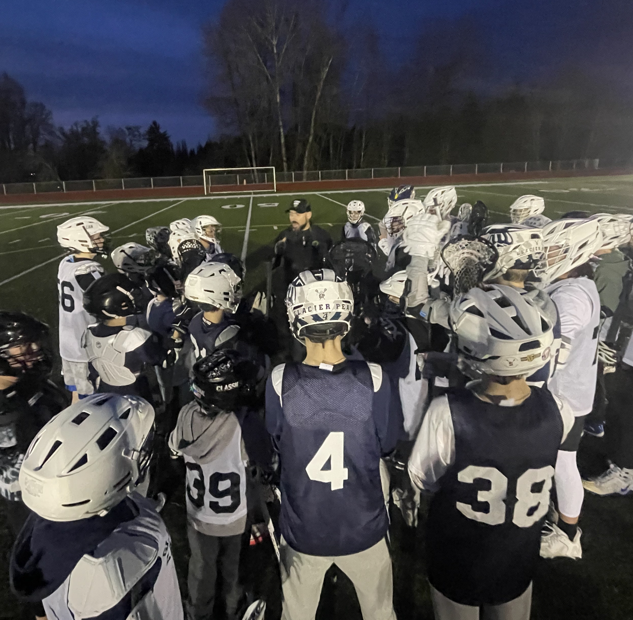 A coach addressing a huddle of young lacrosse players at dusk on a lit field