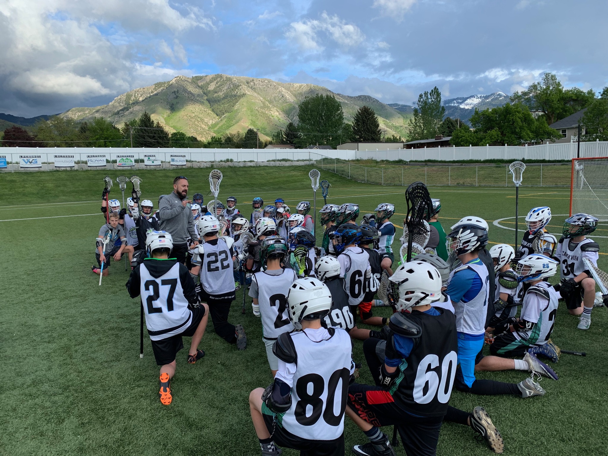A coach addressing a large group of youth lacrosse players kneeling on a field with Utah mountains behind them