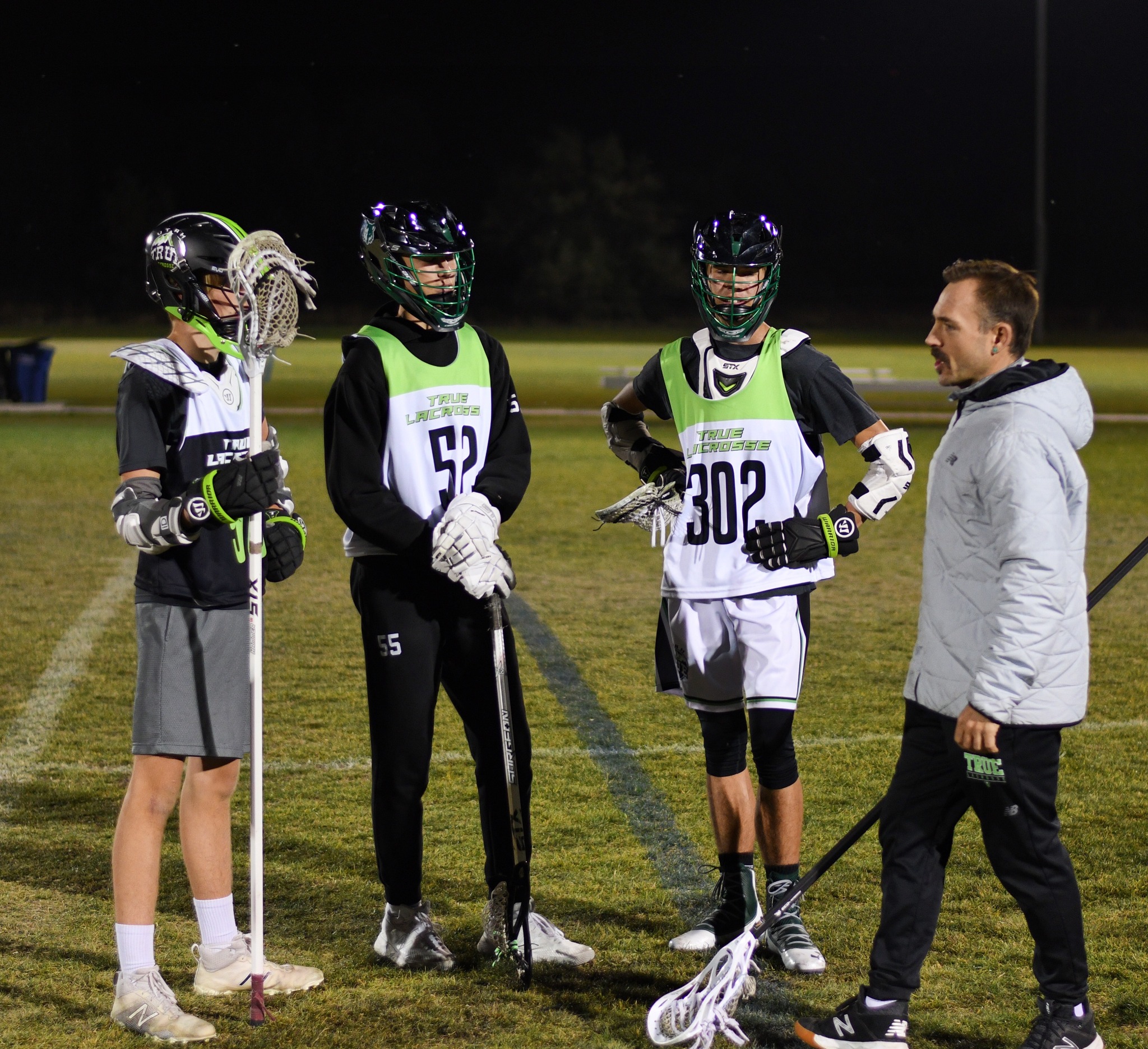 A coach talking with three high-school-aged lacrosse players during an evening practice